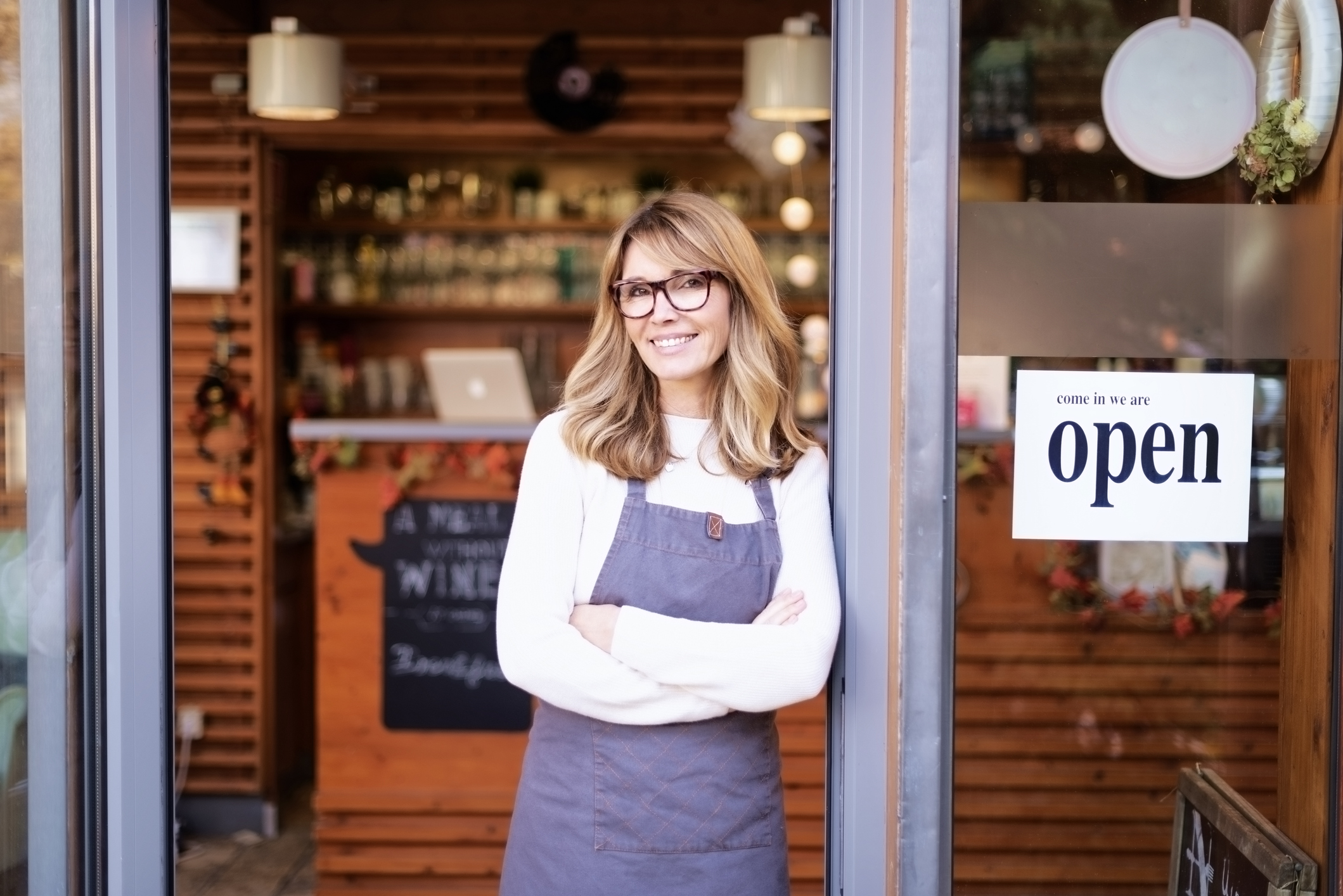 Portrait of confident female owner standing at entrance of cafe. Brunette haired beautiful woman looking at camera and smiling. Open sign in the door.